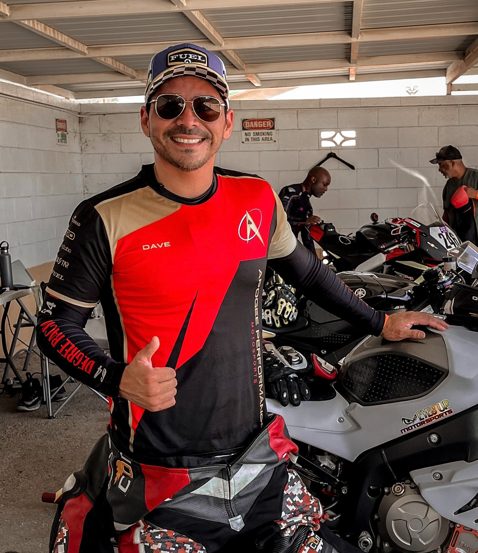 Man in racing gear giving a thumbs up in a garage with motorcycles.